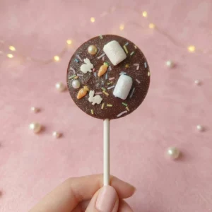 Sucette en chocolat au lait décorée de guimauves, vermicelles colorés et petits motifs sucrés, tenue à la main sur fond rose pastel avec perles décoratives